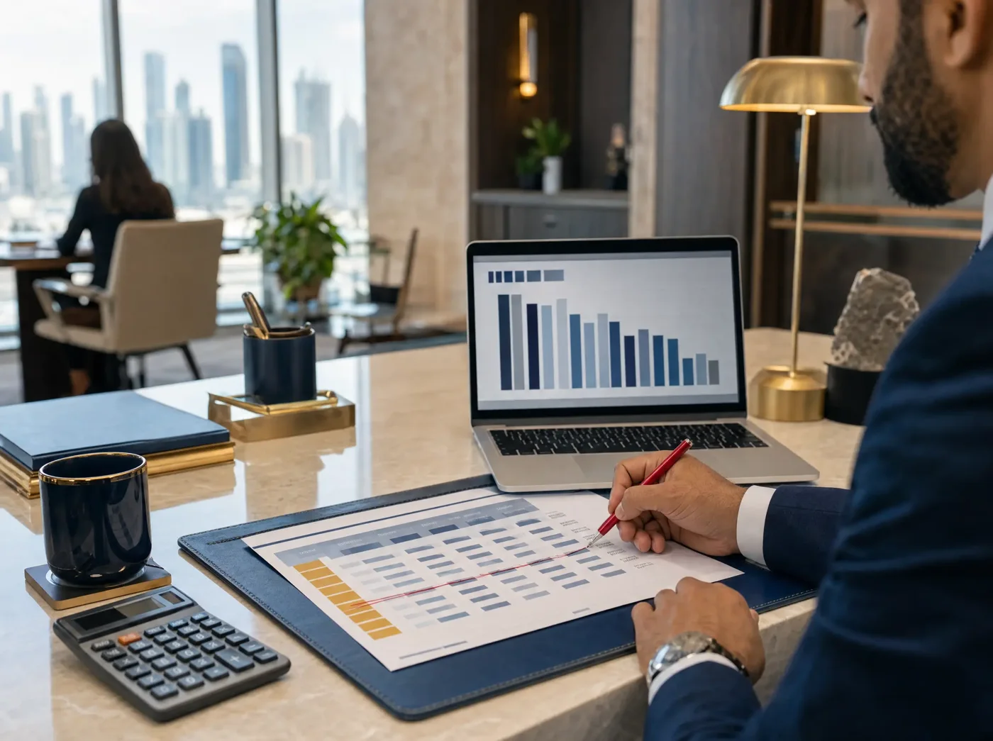 A man in a suit marking up a printed spreadsheet with a red pen at a desk, beside a laptop showing a bar chart and a calculator.