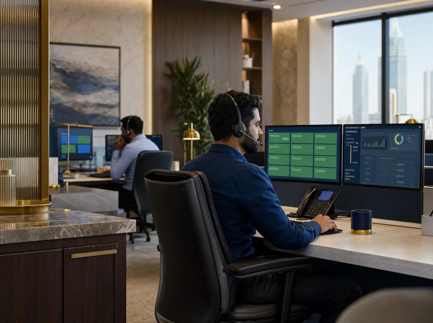 A man wearing a headset at a two-monitor desk showing a green status grid, with another headset worker behind him in an open-plan office.